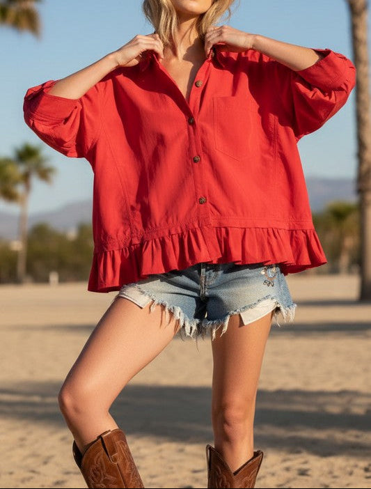 Woman in a red blouse and denim shorts standing on a beach with palm trees in the background.