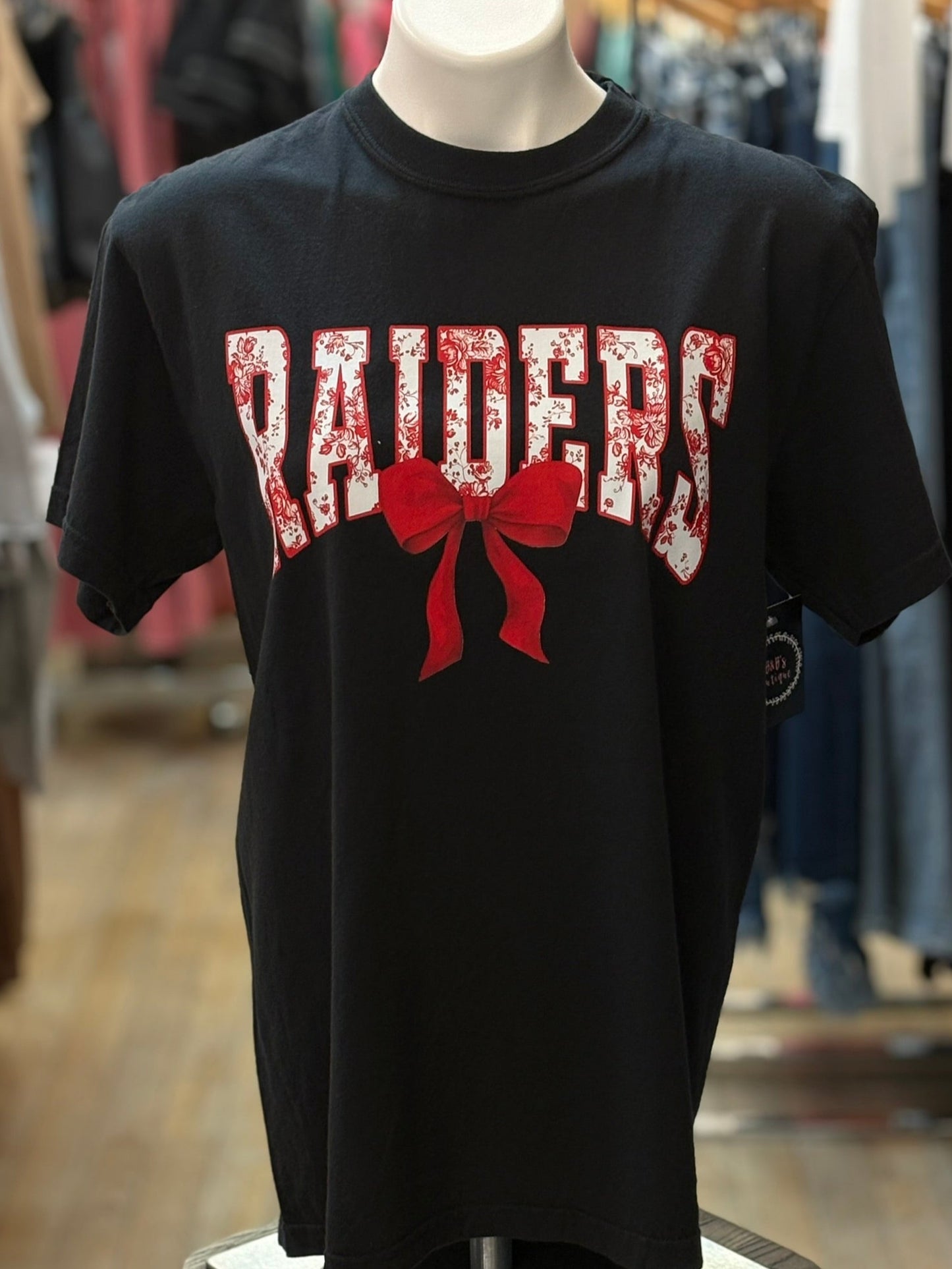 Black t-shirt with 'RAIDERS' and a red bow graphic on a mannequin in a store setting.