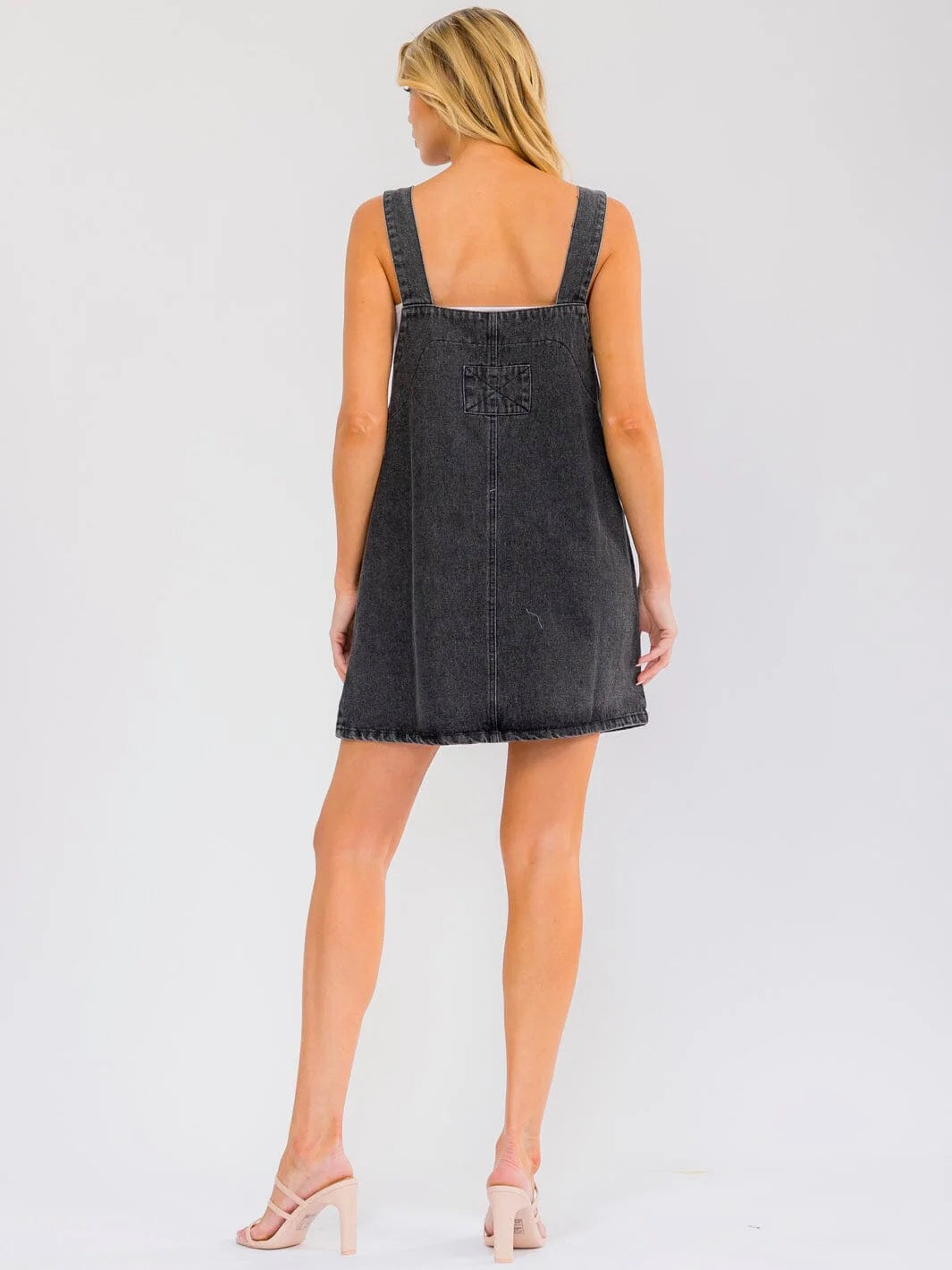 A woman shown from the back wearing a dark gray denim overall dress with frayed hems and shoulder straps, paired with light pink high-heeled sandals, against a plain white studio background.