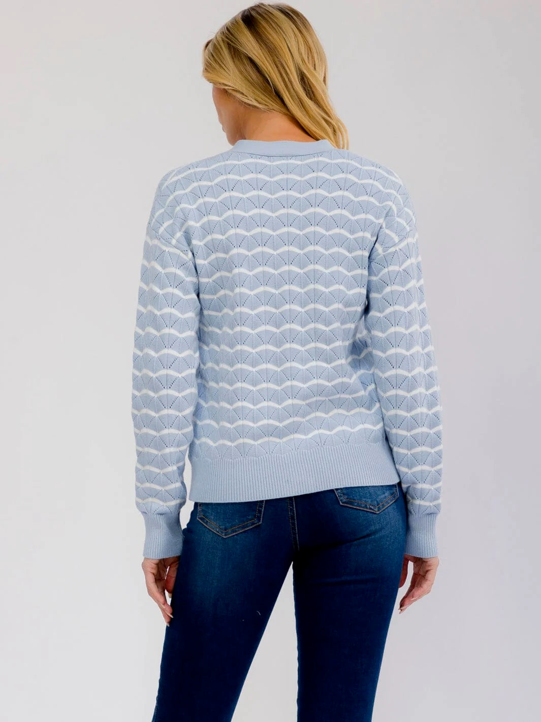 A woman viewed from the back wearing a light blue knit sweater with a white wavy geometric pattern and dark blue jeans, set against a plain light gray studio background.