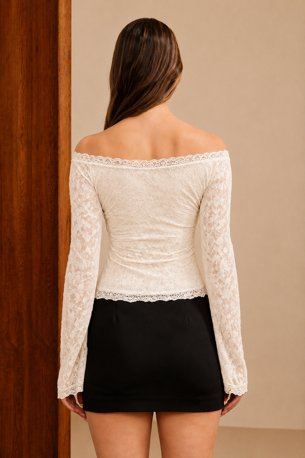 A woman shown from the back wearing a long-sleeve off-the-shoulder lace top in cream and a black mini skirt, standing against a beige wall next to a wooden door frame.