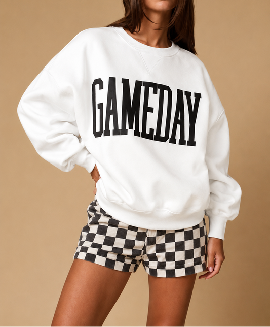 A woman modeling a white crewneck sweatshirt with 'GAMEDAY' printed in large black letters across the chest, paired with black and white checkered shorts, against a solid beige studio background.