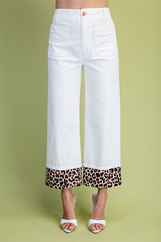 White cropped wide-leg pants with a leopard print hem, modeled with white heeled sandals against a solid light green background.