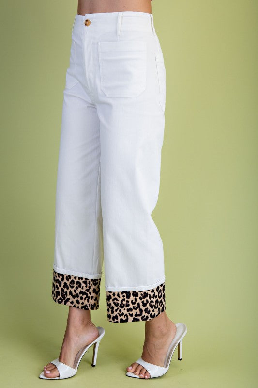 White cropped jeans with a leopard print cuff hem, modeled with white heeled sandals against a light olive green background.
