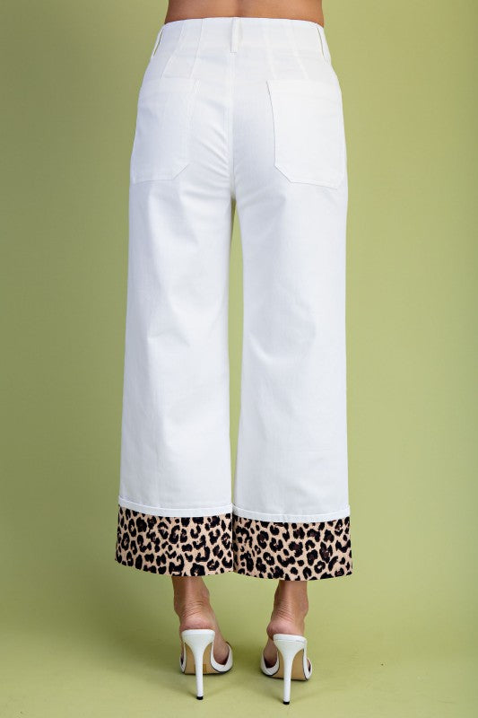 White cropped pants with a leopard-print hem, shown from the back on a model wearing white high heels against a solid olive green background.
