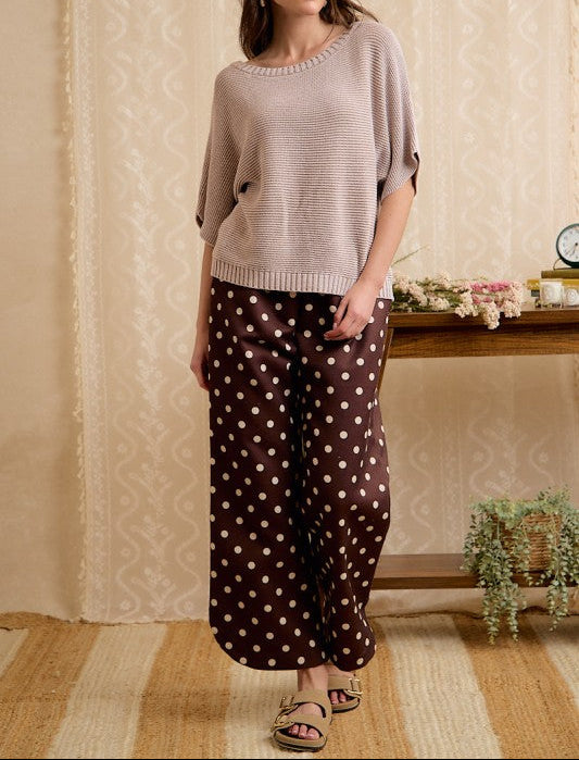 A woman modeling a two-piece outfit consisting of a light beige knit short-sleeve top and dark brown wide-leg pants with white polka dots, standing in a decorated room with patterned wallpaper and a wooden console table.