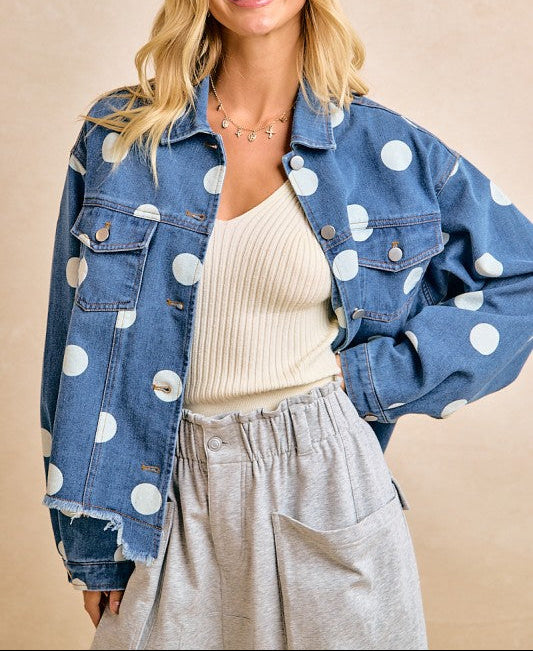 Woman wearing a denim jacket with white polka dots, beige top, and gray pants on a beige background