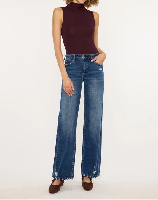 A model with curly brown hair stands against a white studio background wearing a sleeveless dark burgundy mock-neck top and high-waisted, wide-leg blue jeans with brown flat shoes.
