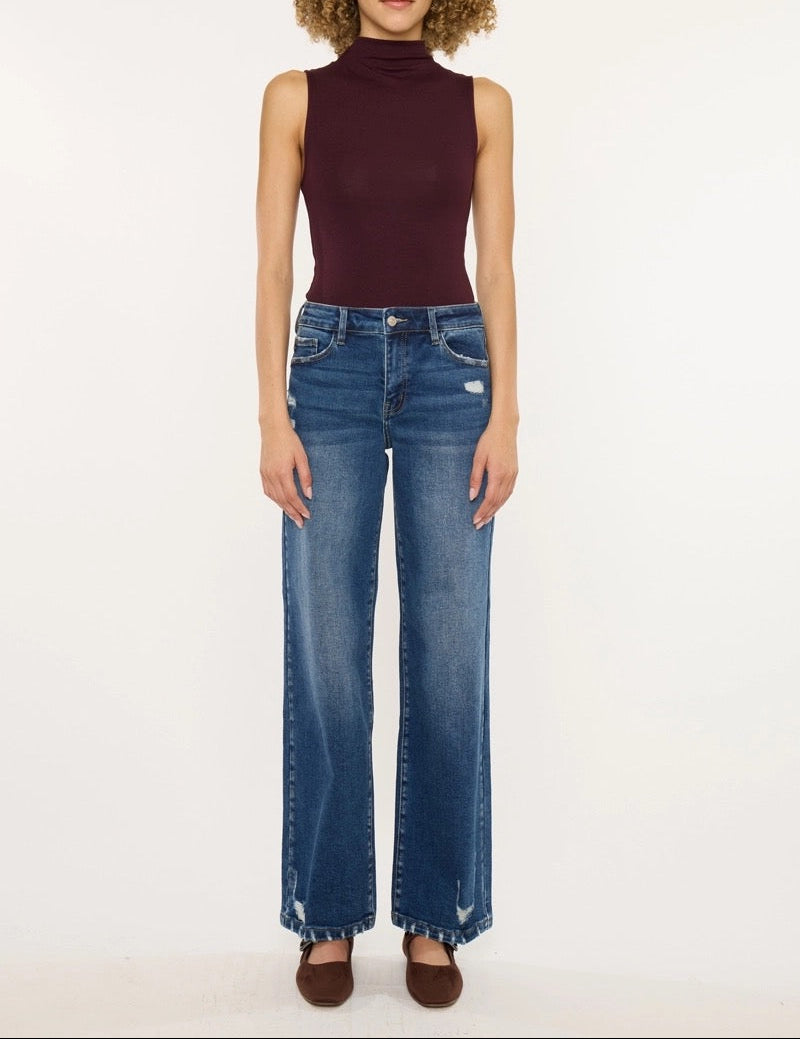 A model with curly brown hair stands against a white studio background wearing a sleeveless dark burgundy mock-neck top and blue wide-leg jeans.