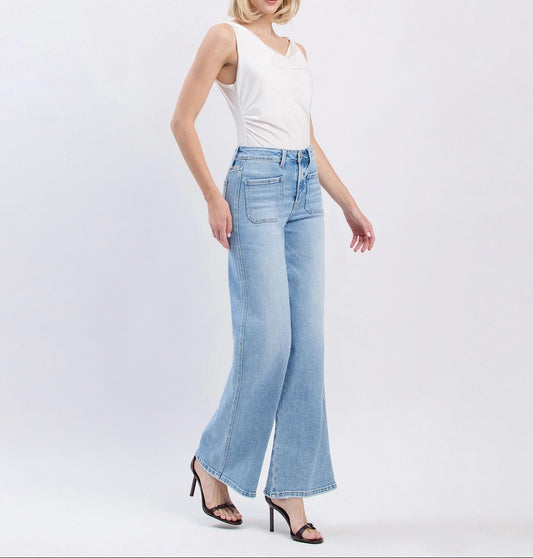 A woman modeling light-wash blue wide-leg jeans with a white sleeveless top and black heeled sandals, standing against a plain light gray studio background.