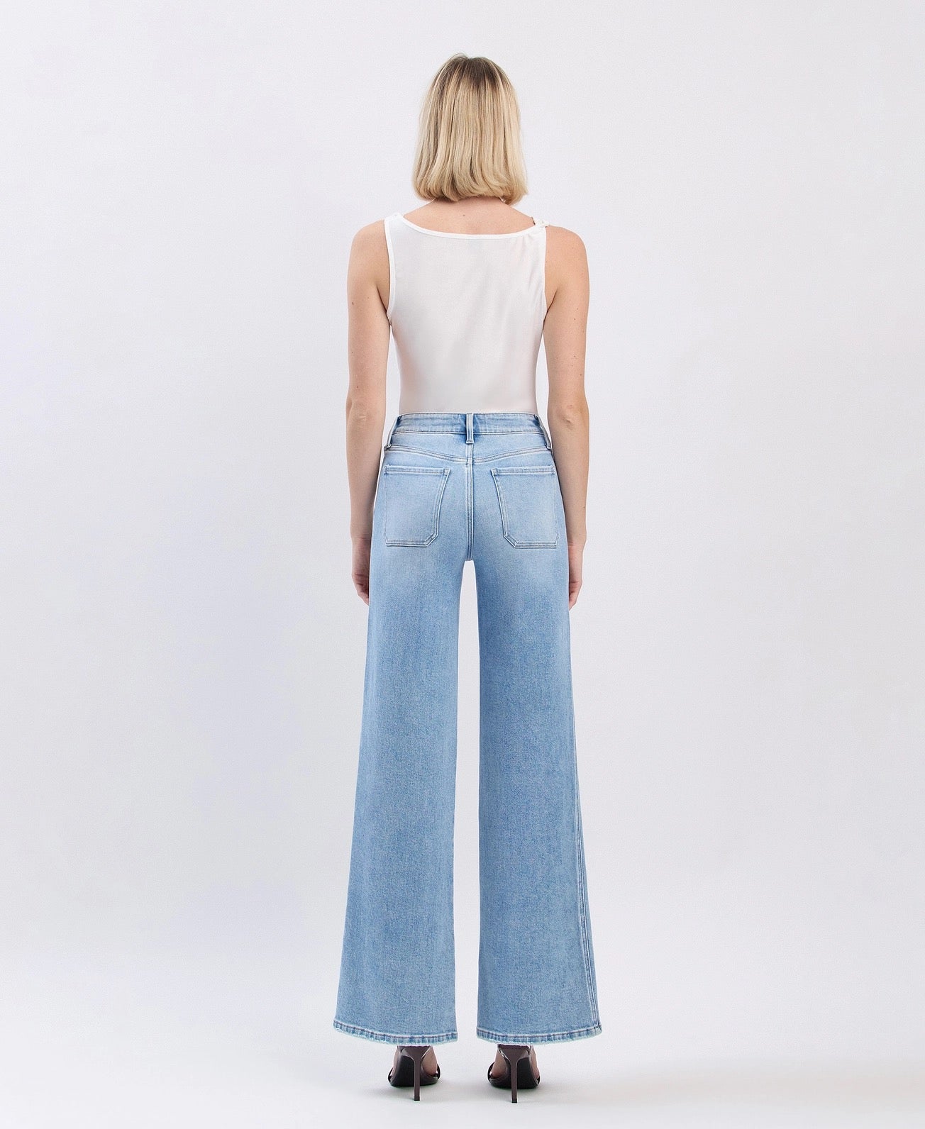 A woman viewed from the back wearing light-wash wide-leg denim jeans and a white sleeveless top, standing against a plain light gray studio background.