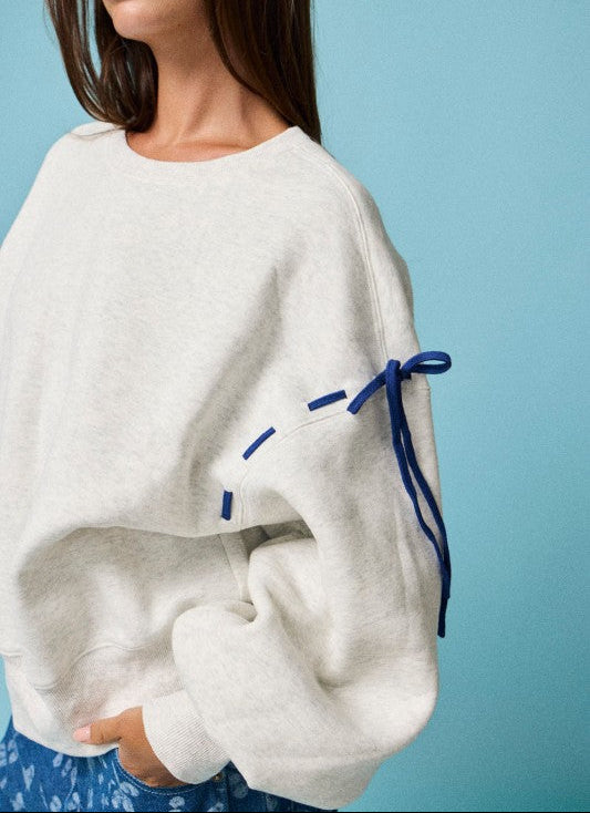 A woman wearing a cream-colored knit sweater with blue ribbon details tied to the sleeve, set against a solid light blue background.