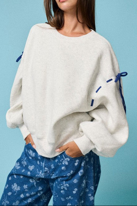 A woman modeling a cream-colored oversized knit sweater with blue drawstring details on the sleeves, paired with blue patterned pants, set against a solid light blue background.
