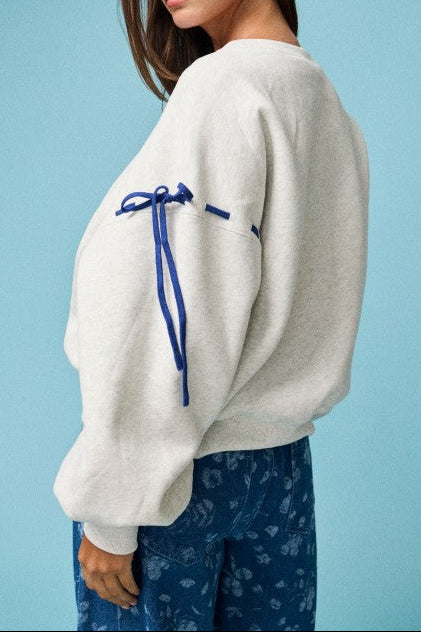 A woman wearing a light heather gray oversized sweatshirt with a contrasting royal blue drawstring detail on the sleeve, paired with blue patterned pants, set against a solid light blue background.