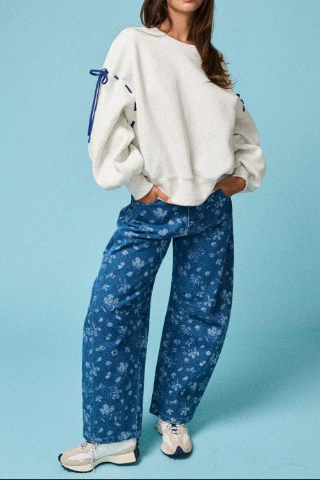A woman modeling a cream-colored oversized sweatshirt with blue drawstrings on the sleeves, paired with blue floral wide-leg jeans and white sneakers, set against a solid light blue background.
