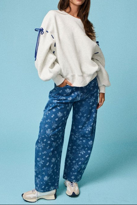 A woman modeling a cream-colored oversized sweatshirt with blue drawstrings and blue patterned wide-leg jeans, standing against a solid light blue background.