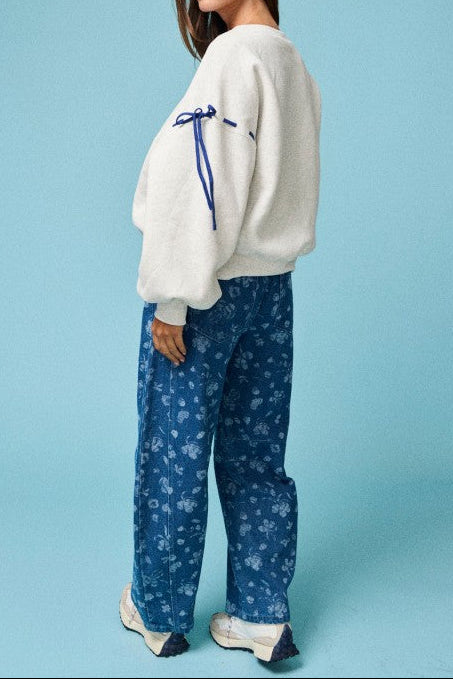 A woman stands against a light blue studio background wearing a cream fuzzy cropped sweater with a blue ribbon detail on the sleeve, blue floral print wide-leg pants, and white sneakers.