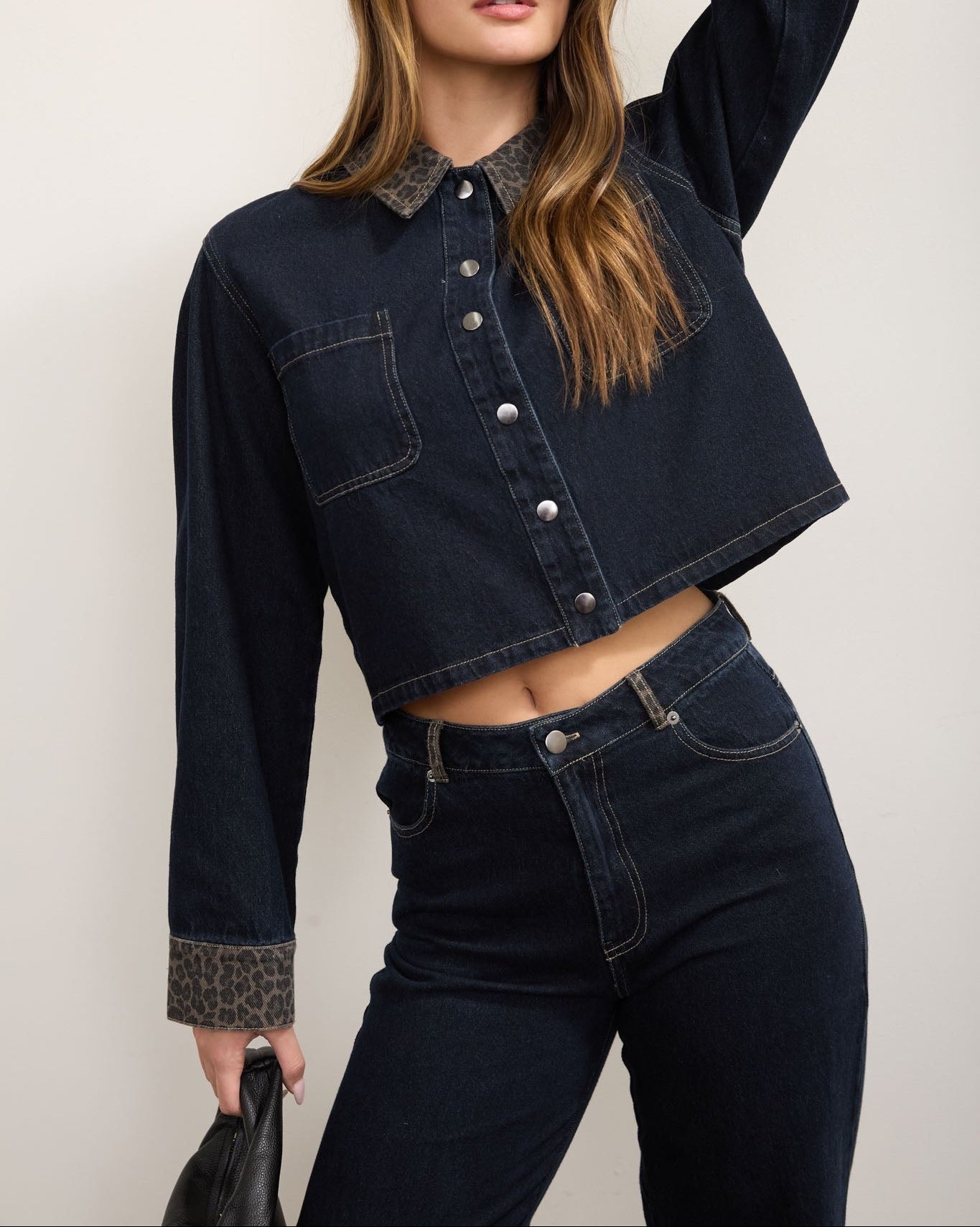A woman modeling a dark wash cropped denim jacket with contrasting leopard-print collar and cuffs, paired with matching dark denim jeans, sunglasses, and a black clutch against a plain light background.