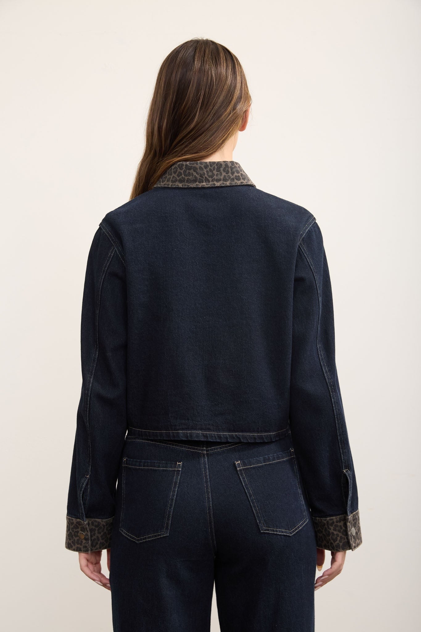 A woman viewed from the back wearing a dark indigo denim jacket with a brown and black patterned knit collar and matching patterned cuffs, paired with dark denim pants, against a plain off-white background.