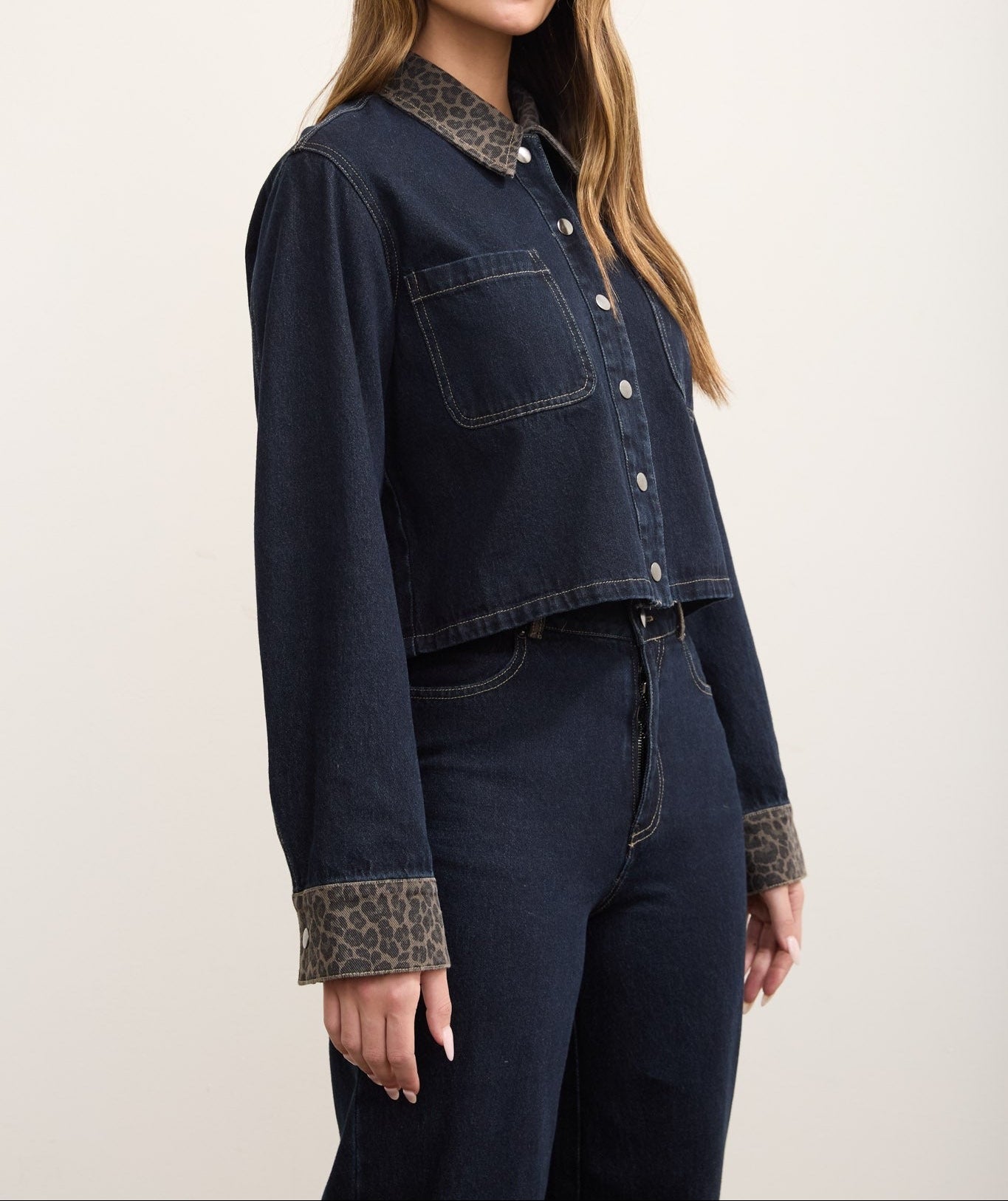 A woman modeling a dark indigo denim jacket with contrasting leopard-print trim on the collar and cuffs, paired with matching dark denim pants against a plain light background.