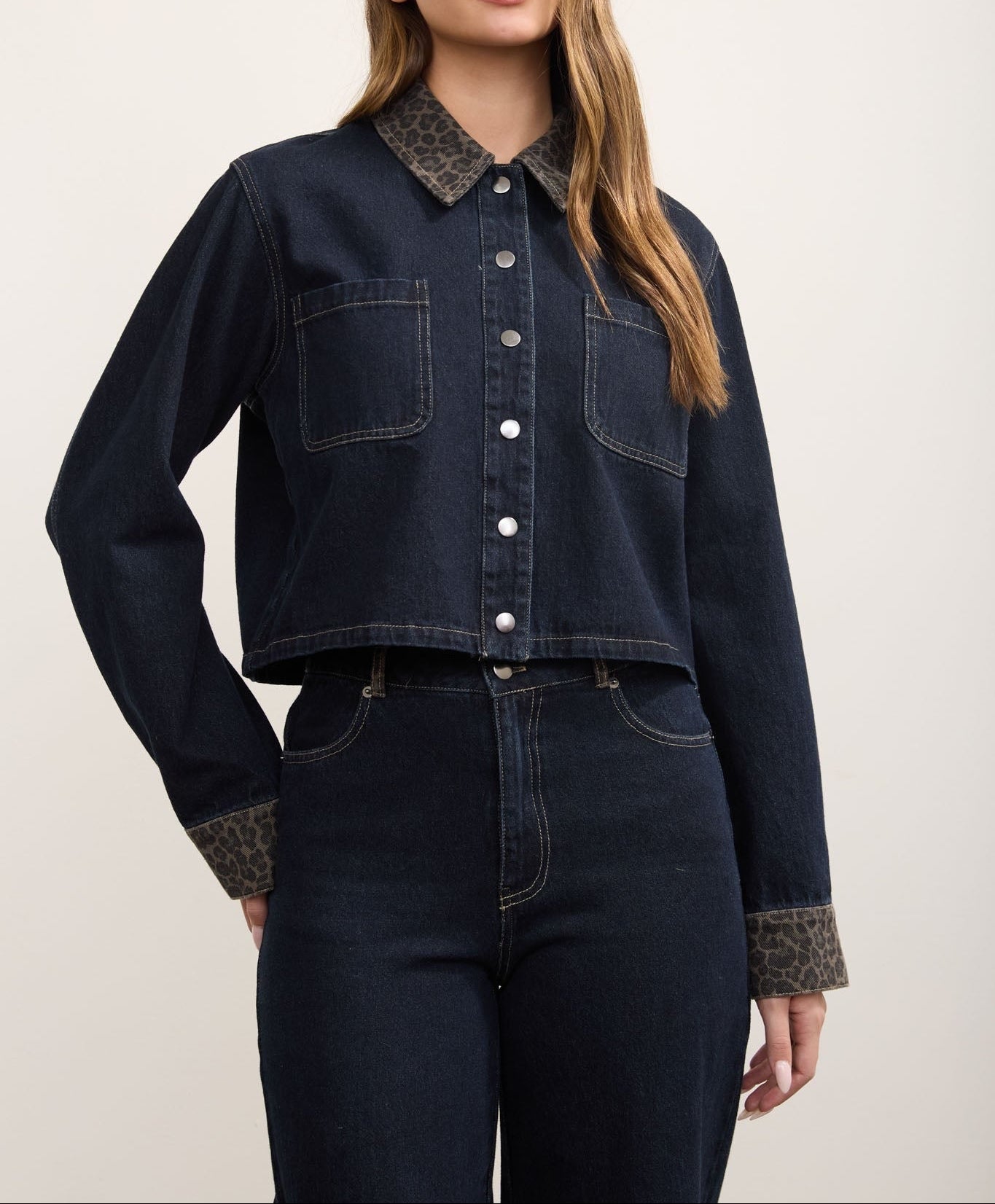 A woman modeling a dark indigo cropped denim jacket with a contrasting leopard-print collar and cuffs, paired with dark jeans against a plain light beige background.