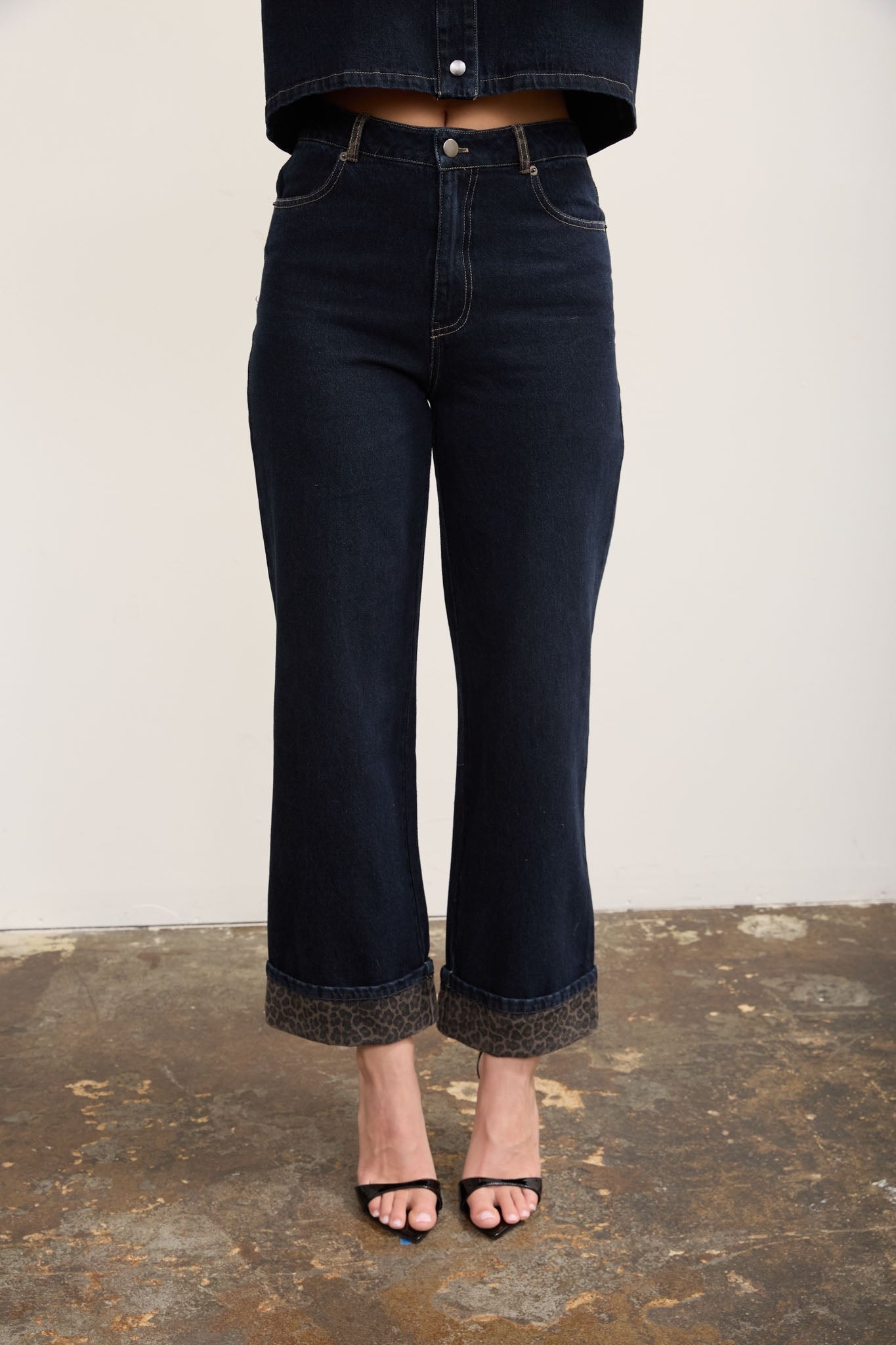 A person wearing dark indigo cropped flare jeans with rolled cuffs and a textured hem, paired with black heeled sandals against a plain light wall and rustic floor.