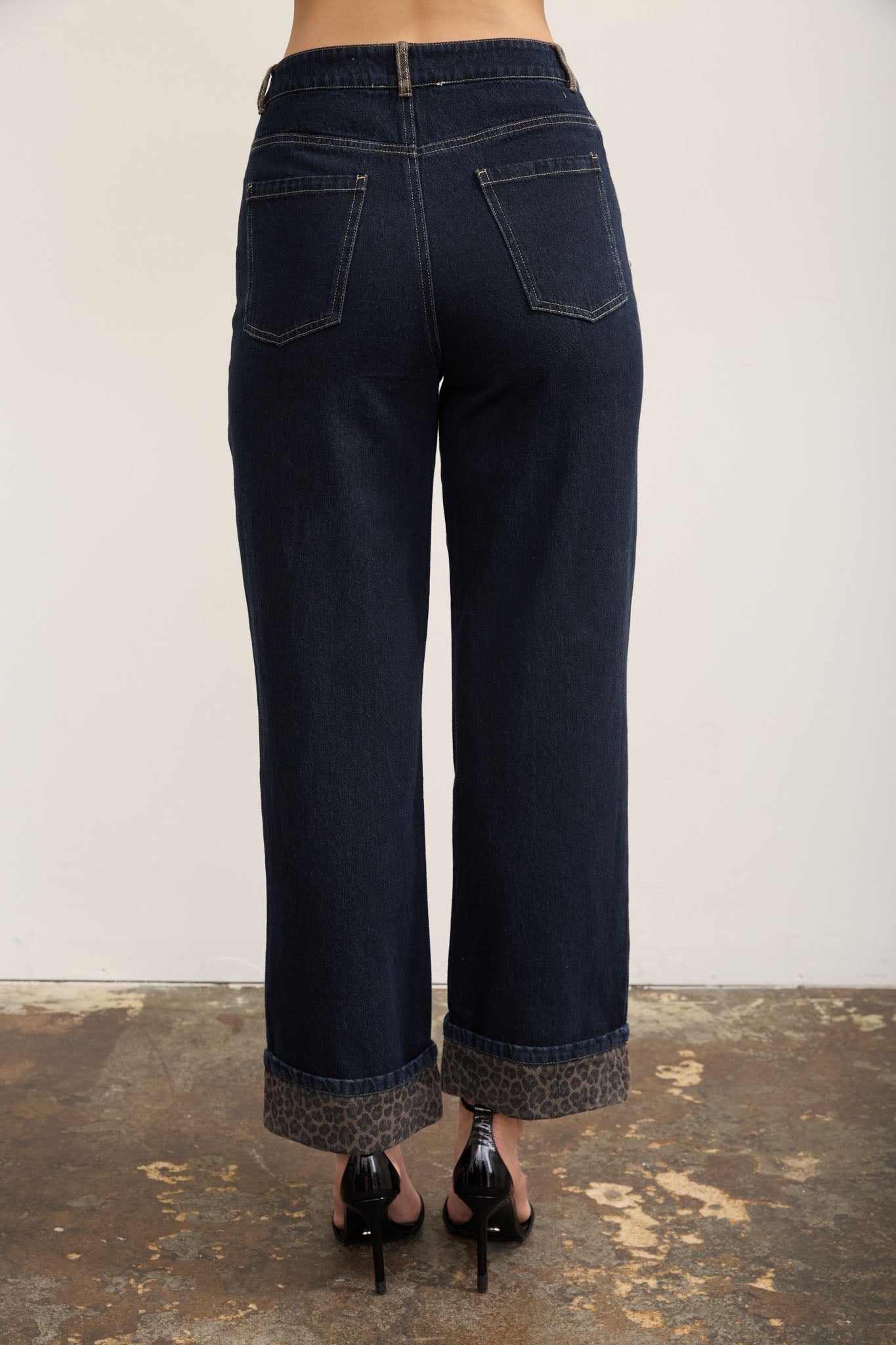 Back view of a person wearing dark blue wide-leg cropped jeans with rolled cuffs and black high heels, standing against a plain off-white wall and textured concrete floor.