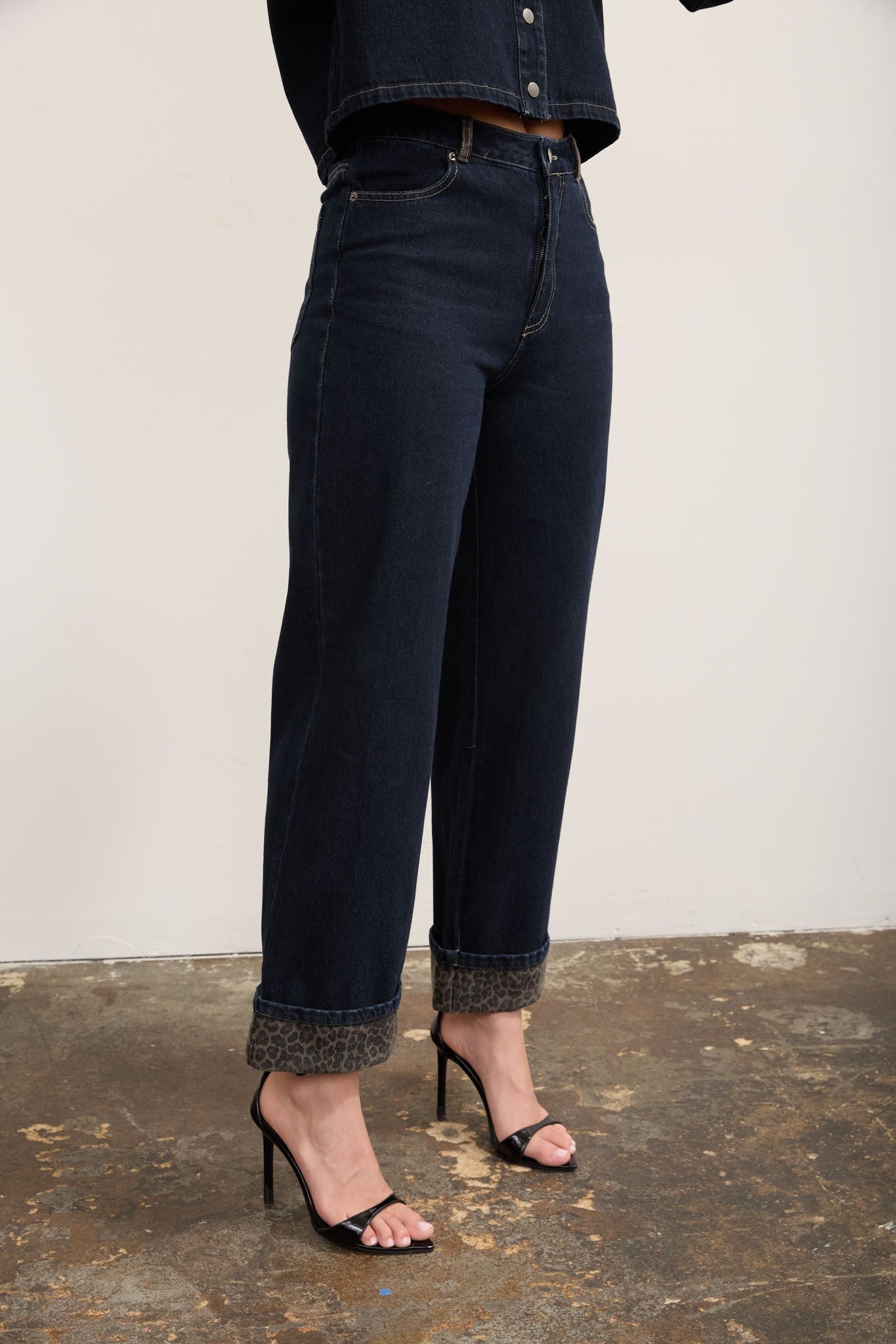 A person wearing dark blue straight-leg denim jeans with rolled cuffs, paired with black high-heeled sandals, shown from the waist down against a plain off-white wall and textured floor.