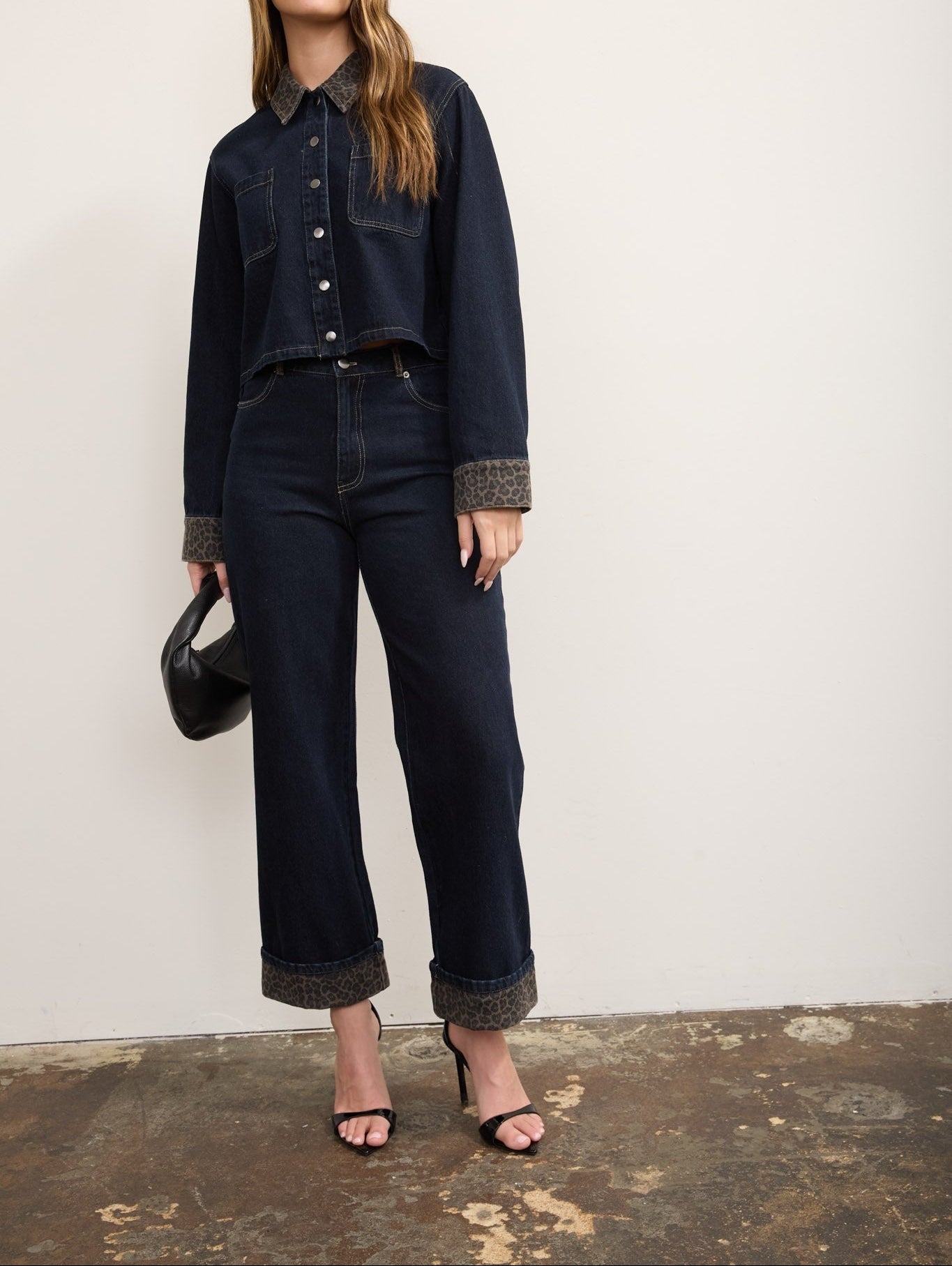 A model wearing a dark wash denim jacket and matching wide-leg jeans with contrasting brown textured cuffs and hem, styled with black sunglasses, a black handbag, and black heeled sandals against a plain off-white wall.