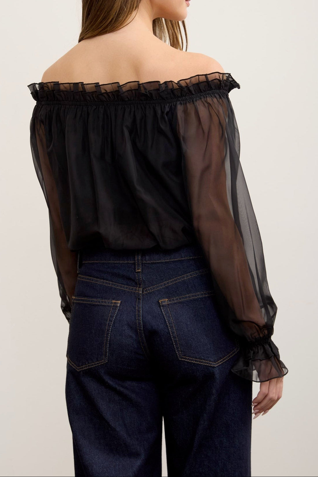 A woman shown from the back wearing a sheer black off-the-shoulder long-sleeve blouse and dark blue jeans against a plain light background.