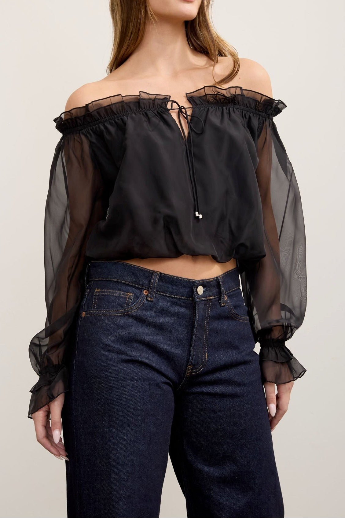 A woman modeling an off-the-shoulder black sheer mesh crop top with ruffled trim and long balloon sleeves, paired with dark blue jeans, against a plain light background.