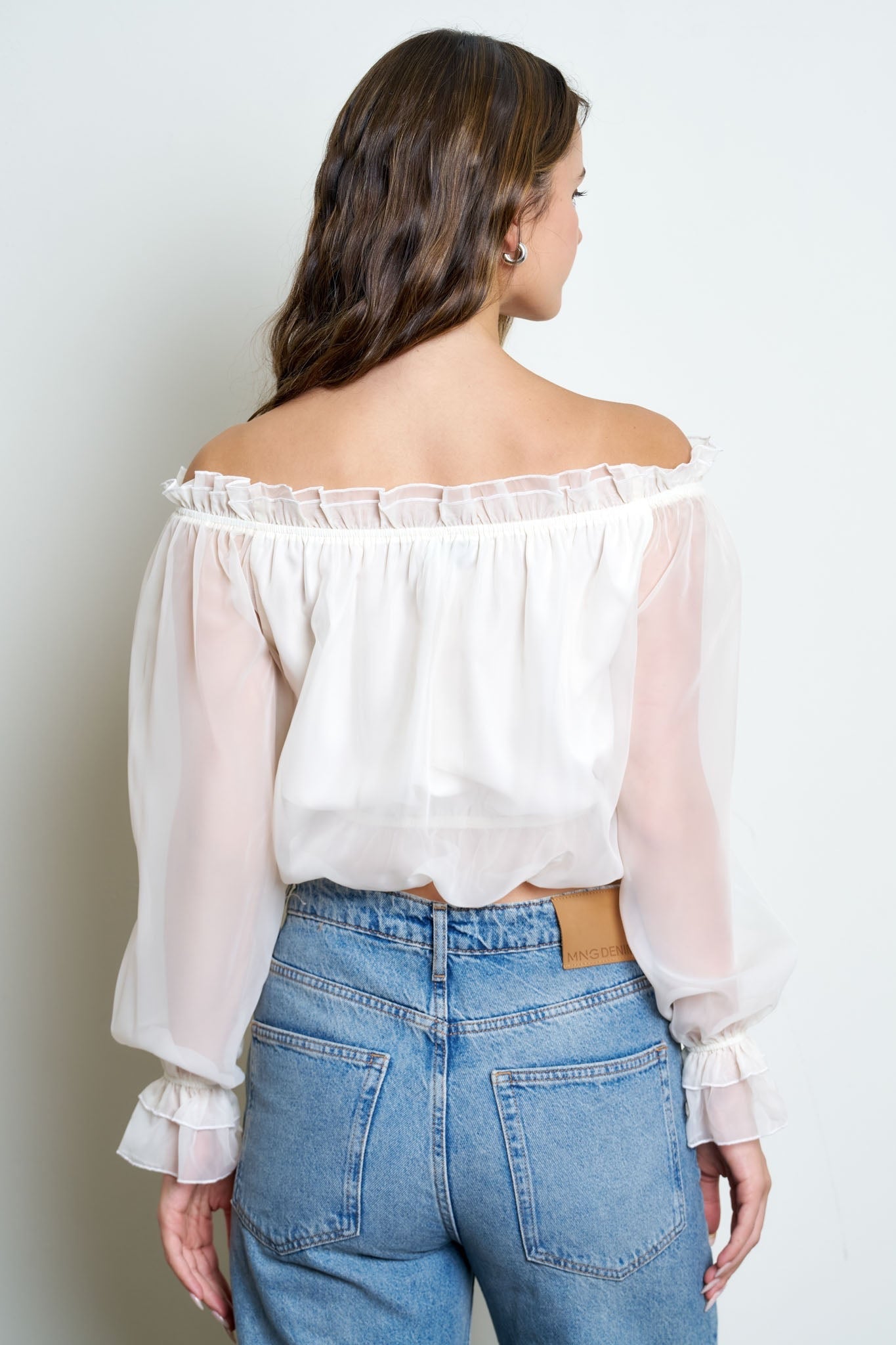 A woman shown from the back wearing a white off-the-shoulder blouse with sheer long sleeves and ruffled trim, paired with light blue jeans, against a plain light gray background.