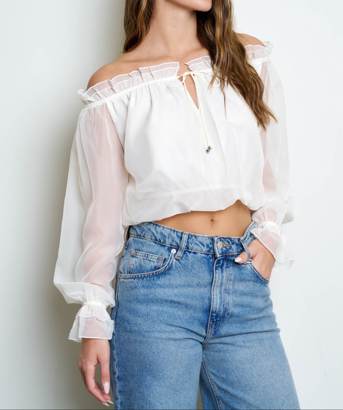 A woman wearing a white off-the-shoulder long-sleeve blouse with ruffled trim and a tie neckline, paired with high-waisted blue jeans, standing against a light gray studio background.