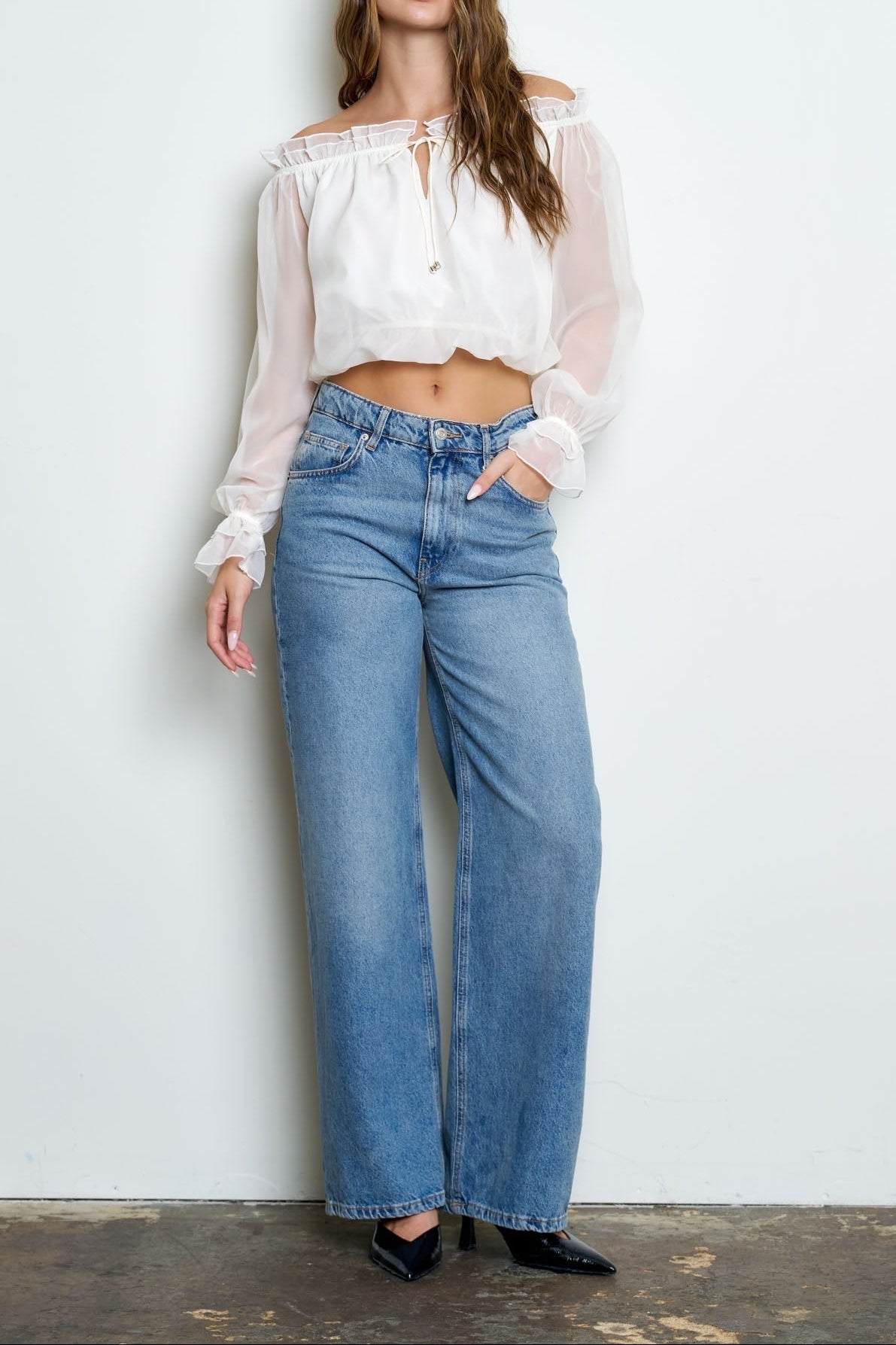 A woman modeling a white off-the-shoulder sheer long-sleeve crop top paired with high-waisted, wide-leg blue jeans and black pointed-toe heels, standing against a plain white wall.