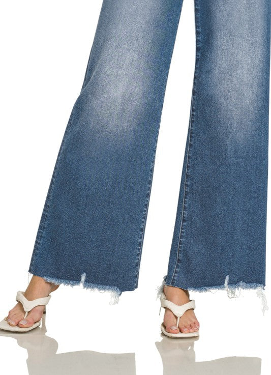 Close-up of a person wearing wide-leg blue denim jeans with a raw frayed hem and white thong sandals, shown against a white studio background.