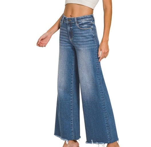 A model wearing a pair of high-waisted, wide-leg blue denim jeans with a frayed hem, shown from the waist down on a plain white background.