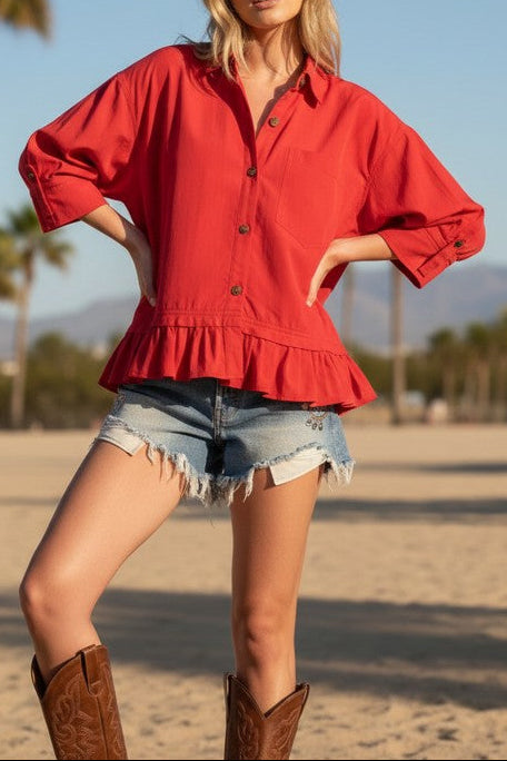 Woman in a red shirt and denim shorts standing on a beach with palm trees in the background.