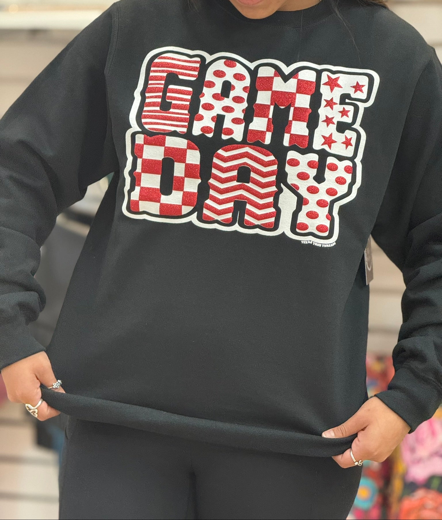 A woman wearing a black crewneck sweatshirt with a large 'GAME DAY' graphic across the chest in red, white, and black patterns, holding the hem of the sweatshirt to show its elasticity.