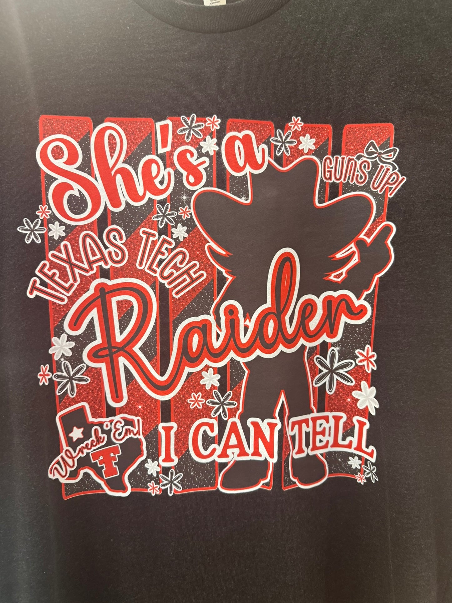 A close-up of a black graphic t-shirt featuring a red and white design with the text 'She's a Texas Tech Raider I Can Tell' and a silhouette of a cowboy.