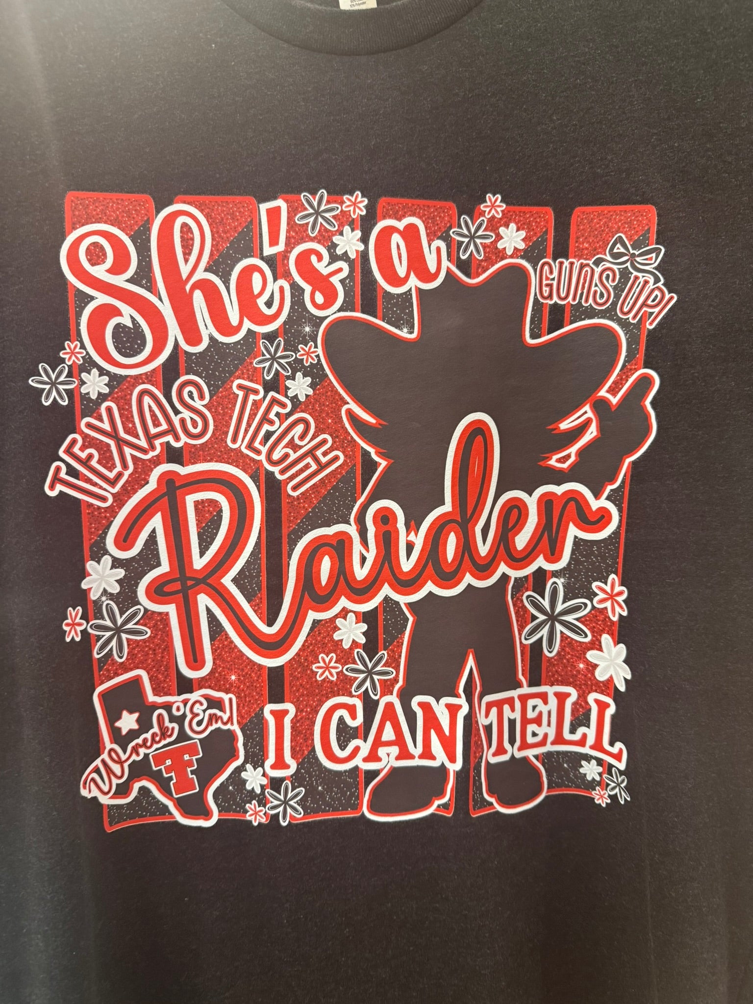 A close-up of a black graphic t-shirt featuring a red and white design with the text 'She's a Texas Tech Raider I Can Tell' and a silhouette of a cowboy.