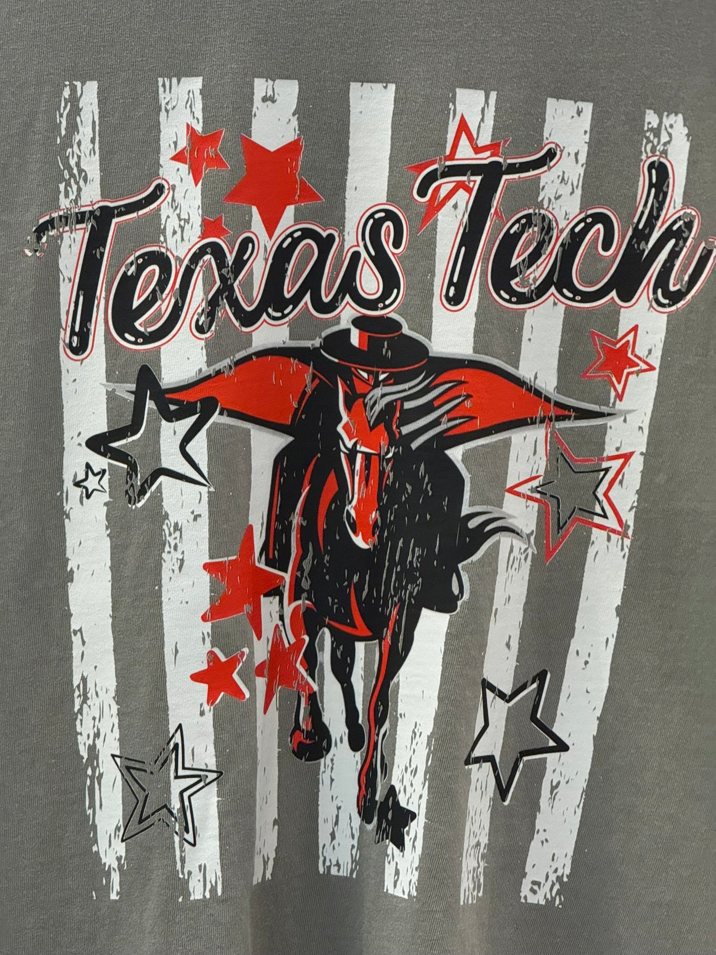Close-up of a gray graphic T-shirt featuring the words 'Texas Tech' in black script over a white striped background, with a cowboy riding a black horse and red and black outlined stars.