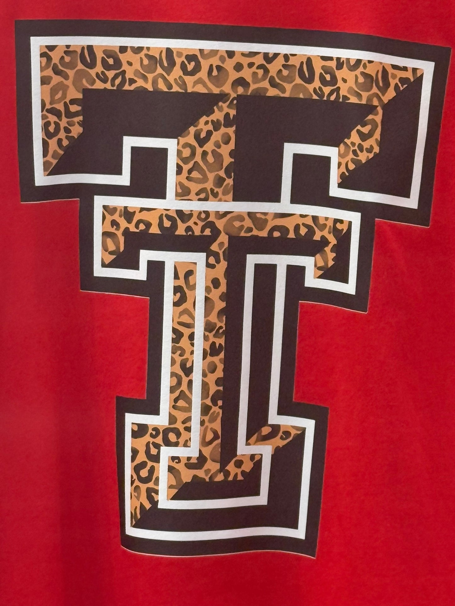 A close-up of a red garment featuring a large collegiate-style letter T graphic with a leopard print fill, dark brown border, and white outline.