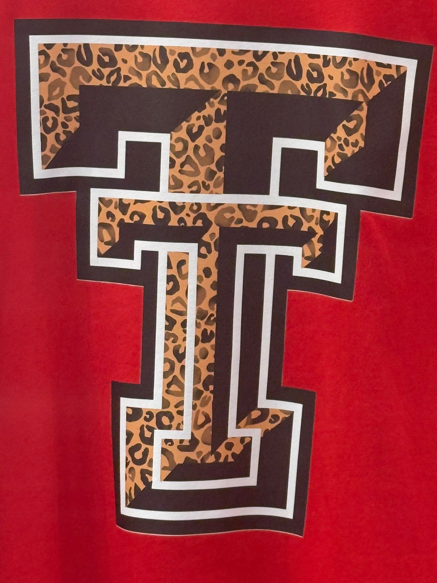 A close-up of a red garment featuring a large collegiate-style letter T graphic with a leopard print fill, dark brown border, and white outline.