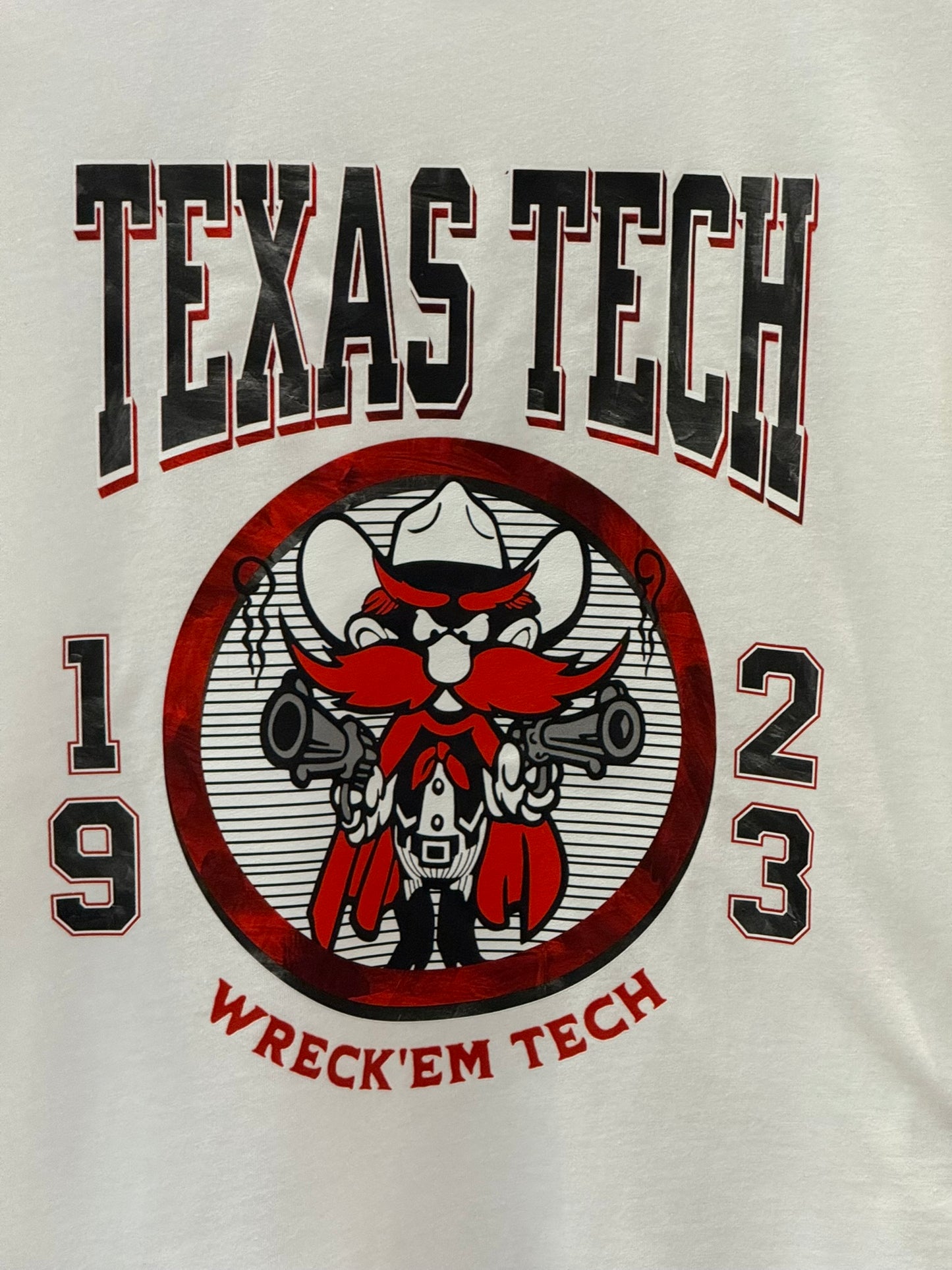 Close-up of a white graphic t-shirt featuring the Texas Tech logo with a red and black cartoon mascot, the text 'TEXAS TECH' and 'WRECK'EM TECH', and the numbers '19' and '23'.