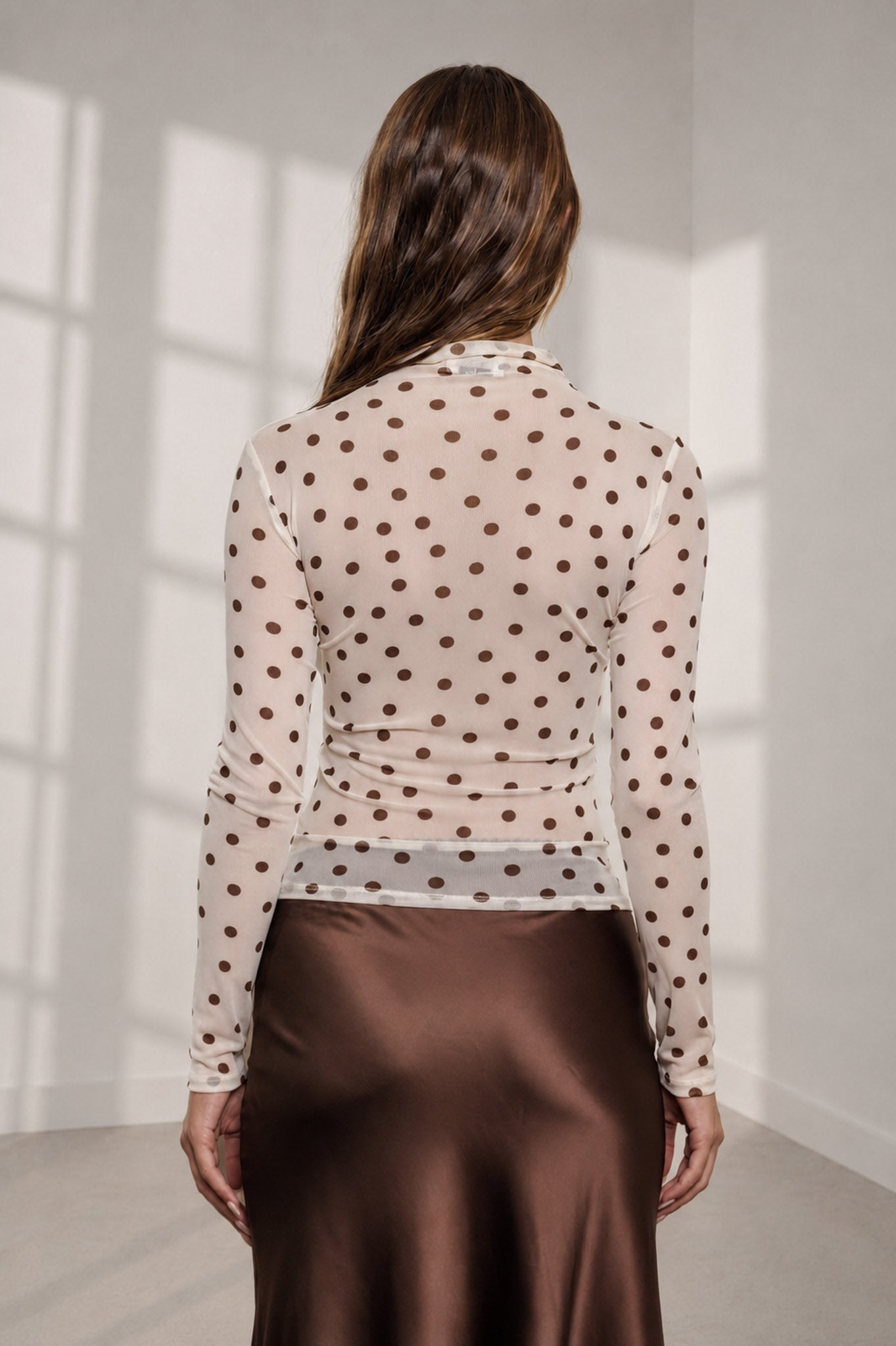 A woman shown from behind wearing a sheer long-sleeve top with a brown polka dot pattern and a brown satin skirt, standing in a brightly lit room with window shadows on the wall.