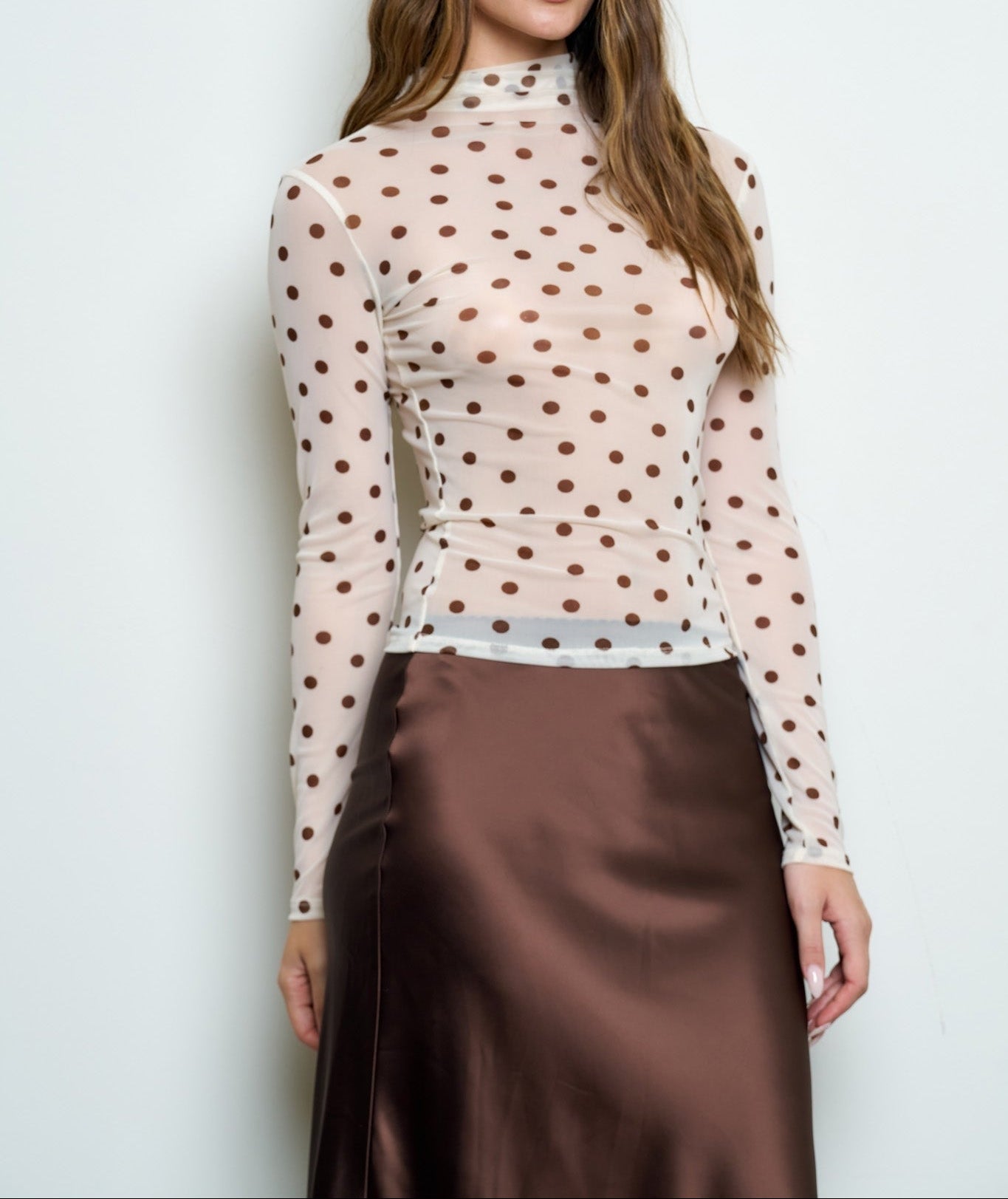 A woman modeling a sheer long-sleeve mock-neck top with a brown polka-dot pattern over a nude base, paired with a brown satin skirt, against a plain light gray background.