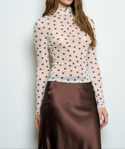 A woman modeling a sheer long-sleeve mock-neck top with a brown polka-dot pattern over a nude base, paired with a brown satin skirt, against a plain light gray background.