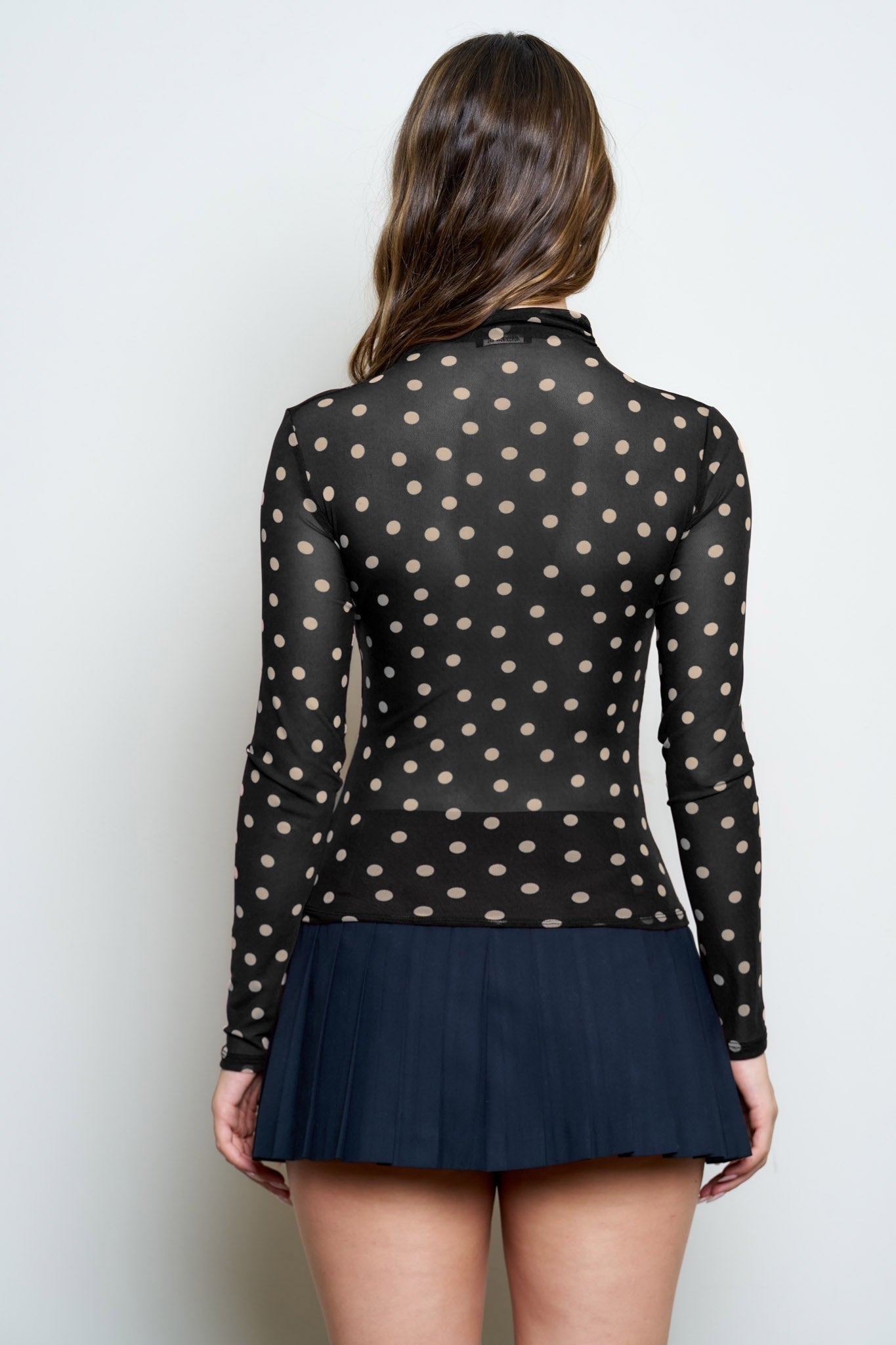 A woman shown from the back wearing a sheer black long-sleeve top with beige polka dots and a dark navy pleated mini skirt, against a plain light gray background.
