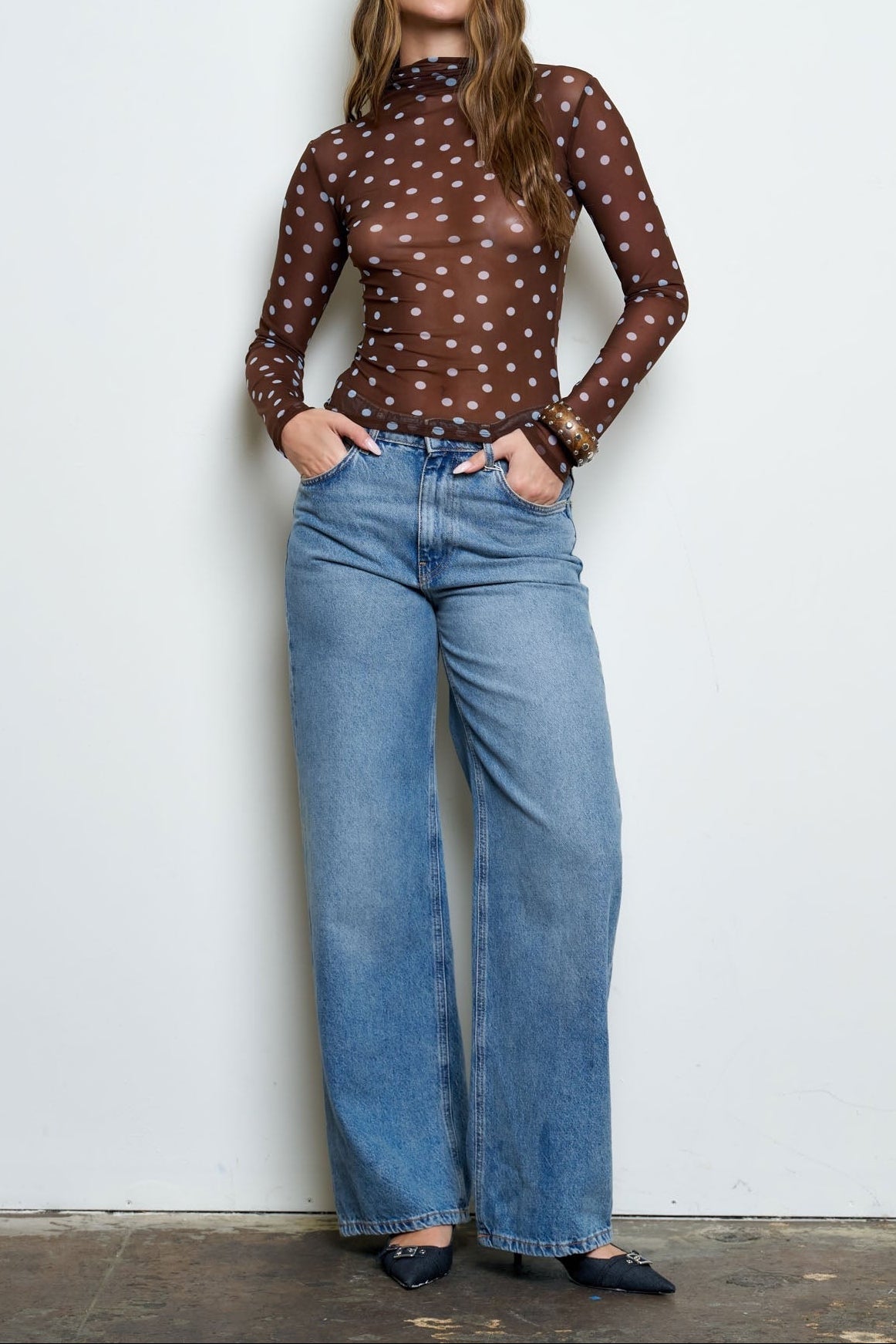A woman stands against a plain white wall wearing a brown sheer long-sleeve top with a white polka-dot pattern, paired with wide-leg light blue jeans and black flats.