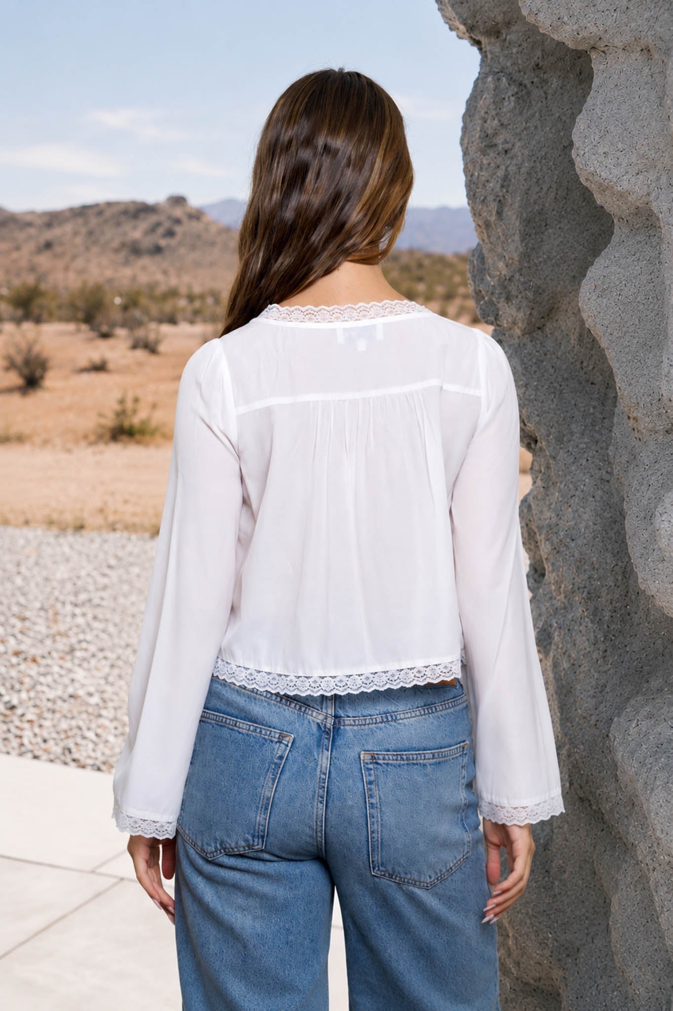 A woman viewed from behind wearing a white long-sleeve blouse with lace trim and blue jeans, standing outdoors next to a large rock with a desert landscape in the background.